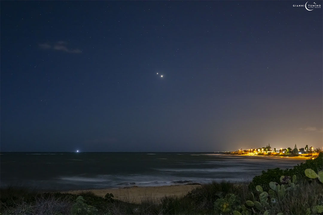 Two bright objects are pictured very near each other in night sky filled with stars. A beach is in the foreground, with some lit structures visible across the water.  Please see the explanation for more detailed information.
